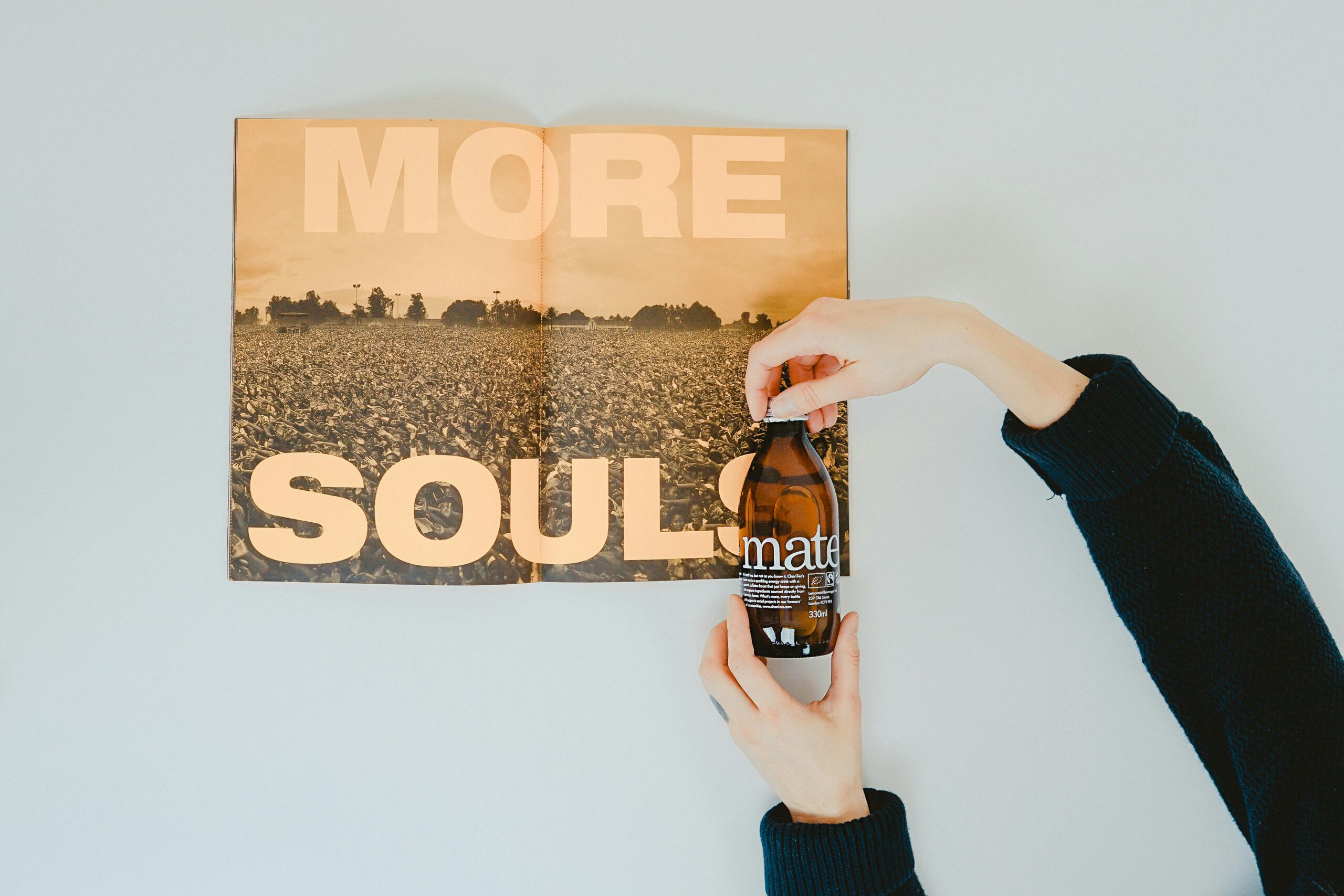 Person holding a brown glass bottle labeled “mate” above an open magazine that reads “MORE SOULS” in large bold letters, symbolizing creativity, authenticity, and the human element behind storytelling and marketing.